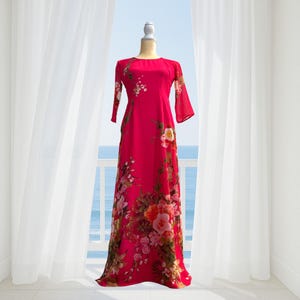 Könnte beinhalten: Ein langes, fließendes, magentafarbenes Kleid mit floralem Muster. Das Kleid hat einen runden Ausschnitt und Dreiviertelärmel. Das florale Design umfasst Rosen und andere Blumen in Rosa-, Gelb- und Grüntönen. Das Kleid wird auf einer Schaufensterpuppe präsentiert.
