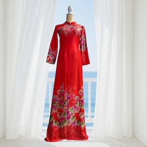 May include: A long, red dress with a mandarin collar and long sleeves. The dress features a floral pattern in shades of pink, purple, and green. The dress is displayed on a mannequin in front of a balcony overlooking the sea.