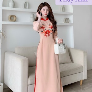 Op de afbeelding: Een vrouw draagt een traditionele Vietnamese jurk, genaamd "Ao Dai", in roze en rood met bloemenborduurwerk. Ze houdt een witte handtas vast.