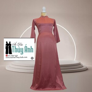 May include: A long, dusty rose-coloured dress with long sleeves and a high collar. The dress has a subtle pattern and is displayed on a mannequin. The dress is from Ao Dai Thuy Anh, as indicated by the logo in the image.