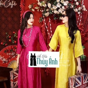 May include: Two women wearing traditional Vietnamese Ao Dai dresses, one in pink and one in yellow, stand in front of a red background with red lanterns and white flowers. The women are holding suitcases with the Union Jack flag.