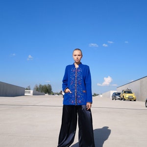 May include: A woman wearing a blue embroidered jacket with a black satin pants and sandals. She is standing in front of a concrete wall and a blue sky.