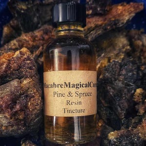 May include: A clear glass bottle with a black cap, labeled "Macabre Magical Cure Pine & Spruce Resin Tincture," sits on a blue speckled plate. Surrounding the bottle are pieces of dark brown and black resin.