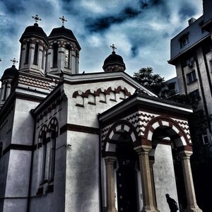 May include: A church exterior with white walls and red brick detailing. The building has several domes, each crowned with a cross. The sky is filled with dark, dramatic clouds. A person is on the steps.