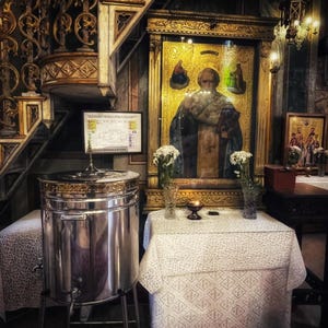 May include: Interior of a church featuring a large, polished metal container on a stand. A table with a white patterned tablecloth and a religious icon are in the background. The walls are decorated with intricate carvings and gold detailing.