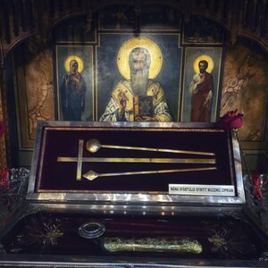 May include: A detailed reliquary featuring gold-coloured liturgical items, such as a spoon, a cross, and a diamond-shaped object, is presented in a glass case. The case is set against a background of religious art and icons. The text "MANA SFANTULUI SFINTIT MUCENIC CIPRIAN" is visible.