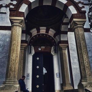 May include: Exterior view of a church with a detailed facade. The building has arched entryways with red brick and white accents. Large columns flank the entrance, and a person is seated on the steps.