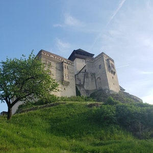 Pode incluir: Um grande castelo claro com várias torres e janelas está no topo de uma colina verde. Uma árvore está no lado esquerdo da imagem, e um céu azul com algumas nuvens está acima do castelo.