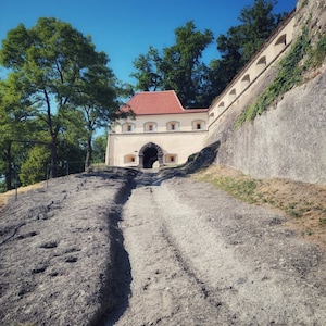 May include: An exterior shot of a stone building with a red tile roof and arched entrance. A stone path leads to the building. The sky is a clear blue, and trees are visible on the left and behind the building.