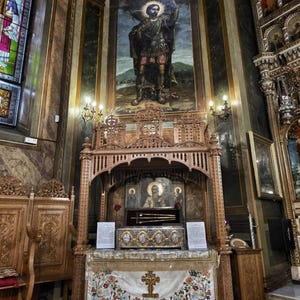 May include: Interior of a church featuring elaborate wooden carvings and religious art. A large mural of a saint is above a decorated altar. The altar is covered with a floral embroidered cloth and a gold cross.