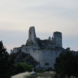Puede incluir: Ruinas de un castillo de piedra sobre una colina, recortadas contra un cielo nublado. Los muros grises del castillo están desgastados, con varias torres y secciones de muro aún en pie. Una persona con un vestido blanco camina por un sendero que conduce al castillo.