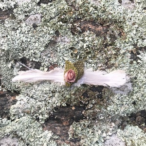 May include: A white fabric headband with a pink fabric rose and green leaves. The headband is lying on a mossy surface.