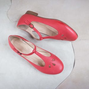 May include: A pair of red leather shoes with a t-strap design and cutouts. The shoes have a low heel and a pointed toe.