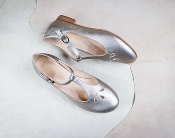 Può includere: Due paia di ballerine in pelle argentata con cinturino a T e dettagli traforati. Le scarpe hanno punta a punta e un tacco basso.