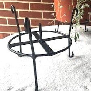 May include: A black metal plant stand featuring a circular frame with a cross-shaped support bar in the center. The stand is supported by four slender legs that curve upward at the top, providing a stable base for holding a potted plant.