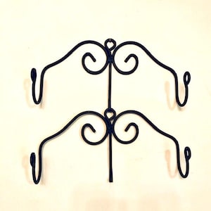 May include: A black metal wall-mounted rack featuring two horizontal tiers. Each tier consists of a central vertical support with symmetrical, curved scrollwork extending outward to end in upward-facing hooks on both the left and right sides.