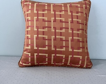 Könnte beinhalten: Ein quadratisches Kissen mit einem geometrischen Muster in Orange- und Goldtönen. Das Kissen hat einen braunen Rand.