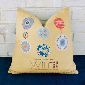 May include: A yellow cushion with a winter theme. It features a string of colourful ornaments with a geometric design. The text "Christmas Ornament Winter Merry Christmas at Home" is printed on the cushion.