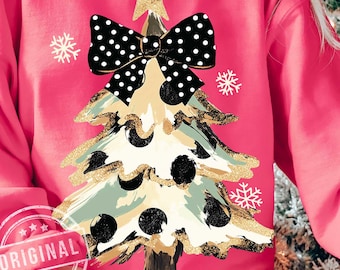 May include: A pink sweatshirt featuring a Christmas tree design. The tree is decorated with black polka dots, gold glitter, and a black bow with white polka dots. A gold star sits atop the tree, and snowflakes are scattered around it.
