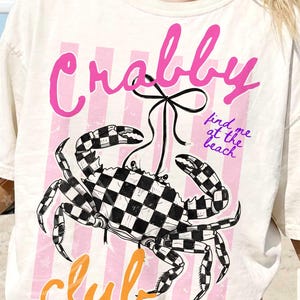 以下が含まれることがあります： クリーム色のTシャツ。白黒の市松模様のカニのグラフィックが特徴です。「Crabby Club」の文字がピンクとオレンジ色でプリントされ、「find me at the beach」というフレーズも含まれています。