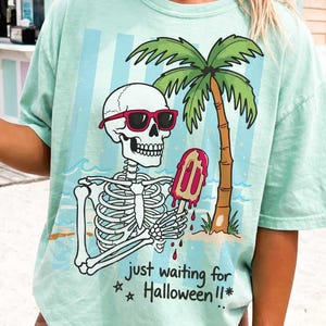 May include: A light green t-shirt featuring a skeleton wearing sunglasses and holding a melting popsicle. A palm tree and the text "just waiting for Halloween!!" are also on the shirt. The shirt has a relaxed fit.