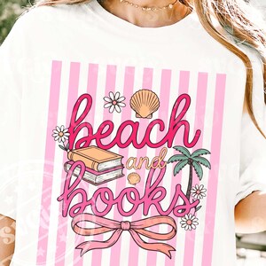 Puede incluir: Camiseta blanca con un dise&ntilde;o de rayas rosas y blancas. La camiseta presenta las palabras "beach and books" en escritura rosa, junto con ilustraciones de una concha, flores, una palmera y una pila de libros.
