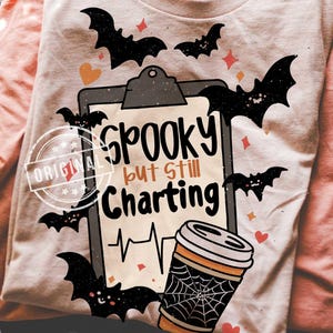 May include: A stack of folded long-sleeved shirts. The top shirt is light beige with a Halloween-themed graphic that includes bats, hearts, and the text "Spooky but still Charting." The graphic also features a coffee cup with a spiderweb design.