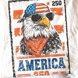 May include: Off-white t-shirt with a graphic of an eagle wearing sunglasses, a bandana, and a headband with the American flag design. The word "AMERICA" is printed in blue and red, with the number "250" above and below the eagle.