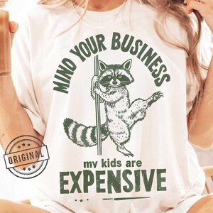May include: White t-shirt with a green graphic of a raccoon holding a pole with the text "Mind Your Business my kids are expensive".