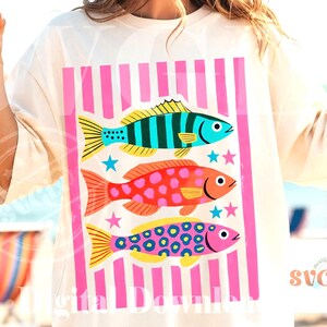 May include: Cream-colored t-shirt featuring a vibrant graphic of three colorful fish with pink vertical stripes. The fish are blue, orange, and pink, with star accents. The shirt is a digital download.
