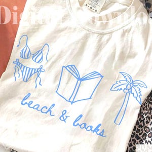 K&ouml;nnte beinhalten: Cremefarbenes T-Shirt mit blauen Illustrationen eines Bikinis, eines offenen Buches und einer Palme. Die Worte "beach & books" sind in blauer Schrift geschrieben. Das Shirt liegt auf Leopardenmuster-Stoff.