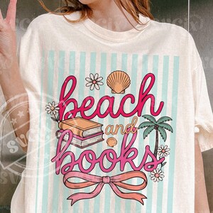 Puede incluir: Camiseta color crema con un gr&aacute;fico colorido. El gr&aacute;fico presenta las palabras "beach and books" en escritura rosa, junto con ilustraciones de una concha, flores, una palmera y un lazo. El fondo tiene rayas azul claro.