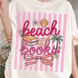 May include: Cream-colored t-shirt featuring a pink and white striped design with the words "beach and books" in pink script. The graphic includes illustrations of books, seashells, flowers, a palm tree, and a bow.