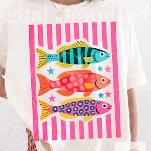 May include: White t-shirt featuring a colorful graphic of three stylized fish. The fish are in shades of blue, orange, and pink, with yellow fins and patterned details. The design includes pink vertical stripes and stars.