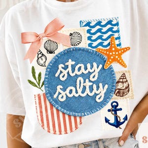 Peut inclure: T-shirt blanc avec un motif patchwork comprenant un n&oelig;ud rose, des coquillages, un cercle bleu avec le texte "stay salty", une &eacute;toile de mer orange et une ancre bleue. Le t-shirt a un th&egrave;me nautique.