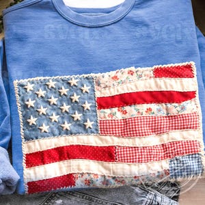 May include: A light blue sweatshirt with a patchwork American flag design. The flag features white stars on a blue background and red and floral print stripes. The sweatshirt is folded on a white surface.