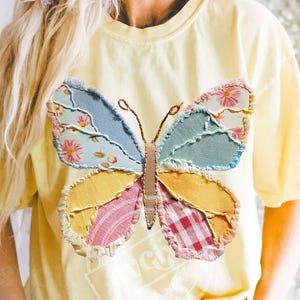 Peut inclure: T-shirt jaune clair orné d'un grand papillon coloré. Les ailes du papillon sont faites de tissu patchwork bleu, jaune, rose et vert, avec des motifs floraux et vichy. Le corps du papillon est marron.