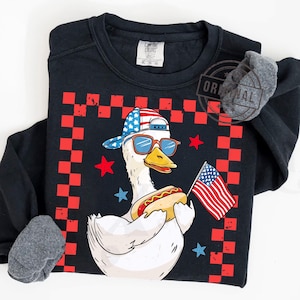 May include: A white duck wearing a red, white, and blue baseball cap and sunglasses, holding a hot dog in one hand and an American flag in the other. The duck is standing on a black and red checkered background.