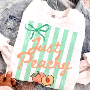 May include: White sweatshirt with green vertical stripes and the text "Just Peachy" in orange script. A green bow and peach graphic are also featured. The sweatshirt is displayed on a denim jacket.