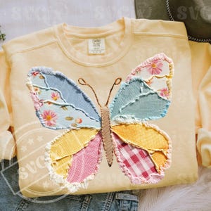 May include: A light yellow sweatshirt featuring a large, colorful butterfly applique. The butterfly wings are made of patchwork fabric in shades of blue, pink, yellow, and green, with floral and gingham patterns. The sweatshirt has a crew neck and the brand name "Comfort Colors" is visible.
