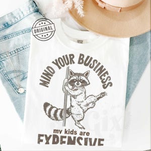 Pu&ograve; includere: T-shirt bianca con grafica di procione che tiene un palo, e il testo "MIND YOUR BUSINESS" e "my kids are FYDENSIVE". La maglietta &egrave; accanto a un cappello beige e jeans blu.