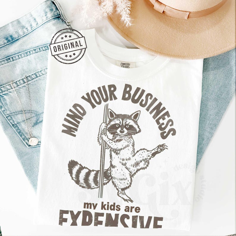 Pu&ograve; includere: T-shirt bianca con grafica di procione che tiene un palo, e il testo "MIND YOUR BUSINESS" e "my kids are FYDENSIVE". La maglietta &egrave; accanto a un cappello beige e jeans blu.