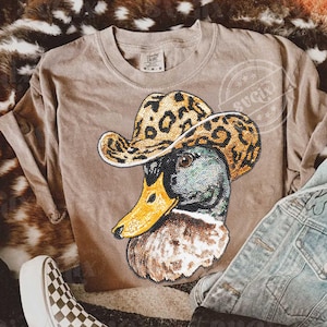 May include: A tan-colored sweatshirt featuring a detailed graphic of a duck wearing a leopard print cowboy hat. The duck's head is a mix of green, brown, and black, with a yellow beak. The shirt has a crew neck and long sleeves.