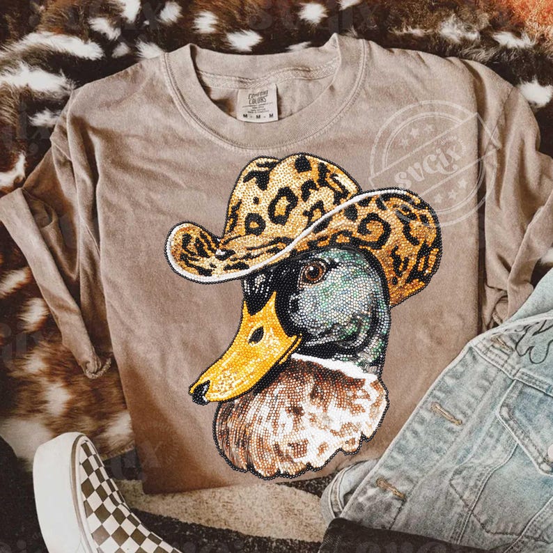 May include: A tan-colored sweatshirt featuring a detailed graphic of a duck wearing a leopard print cowboy hat. The duck's head is a mix of green, brown, and black, with a yellow beak. The shirt has a crew neck and long sleeves.