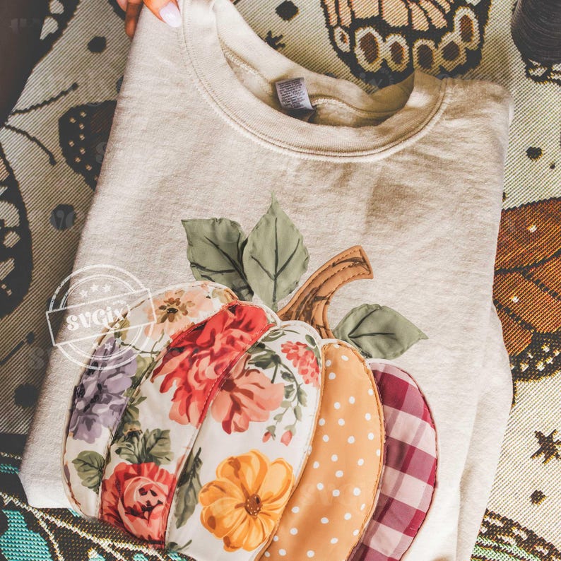 May include: A beige sweatshirt featuring a colorful, appliqued pumpkin design. The pumpkin is composed of fabric patches with floral, polka dot, and plaid patterns. Green leaves and a brown stem complete the autumn-themed design.
