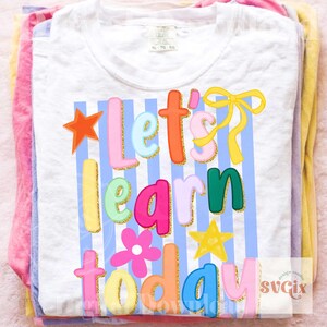 May include: White t-shirt with the phrase "Let's Learn Today" in colorful, playful lettering. The design includes stars, a flower, and a bow, all with gold glitter accents. The shirt is folded with other pastel-colored shirts.