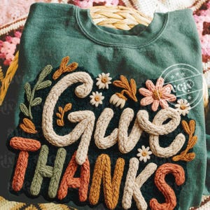 May include: A dark teal sweatshirt with the words "Give Thanks" in a cream-colored, textured font. The design includes floral and leaf accents in shades of orange, green, and pink. The sweatshirt is folded on a woven basket.