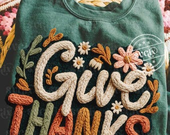 May include: A dark teal sweatshirt with the words "Give Thanks" in a cream-colored, textured font. The design includes floral and leaf accents in shades of orange, green, and pink. The sweatshirt is folded on a woven basket.