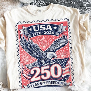 May include: Cream-colored t-shirt featuring a patriotic design. The graphic includes a stylized stamp with the word "USA", the years "1776-2026", an eagle, fireworks, and the text "250 YEARS OF FREEDOM".