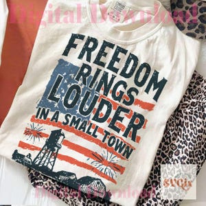 May include: Cream-colored t-shirt with the text "FREEDOM RINGS LOUDER IN A SMALL TOWN" in navy blue. The design incorporates a stylized American flag with red and blue elements, a water tower, and fireworks. The shirt is folded with leopard print pants.